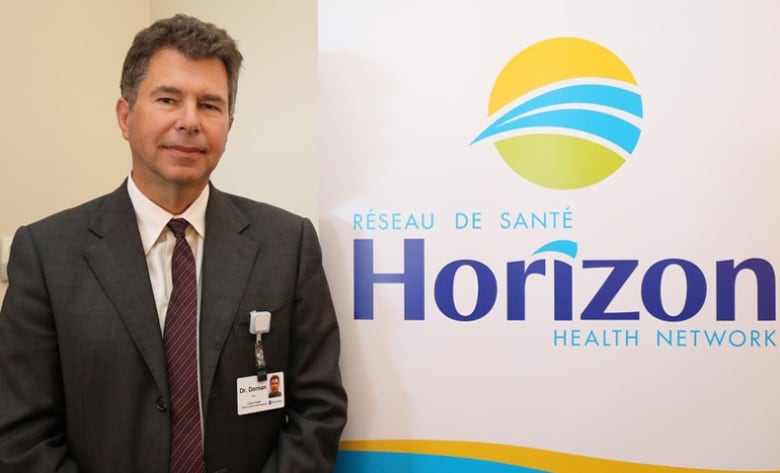 A man wearing a suit and an ID badge on his pocket poses in front of a wall with a large logo for Horizon Health Network. 