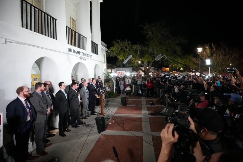 Several people are lined against a wall, at a distance from a large throng of other people who have cameras, boom microphones.